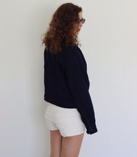 A woman with curly brown hair and sunglasses wearing a dark navy blue cropped denim jacket and white textured shorts, posing against a plain white wall.