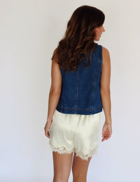 A woman modeling a sleeveless blue denim vest with front tie details over a white top and yellow satin shorts with lace trim, set against a plain background.