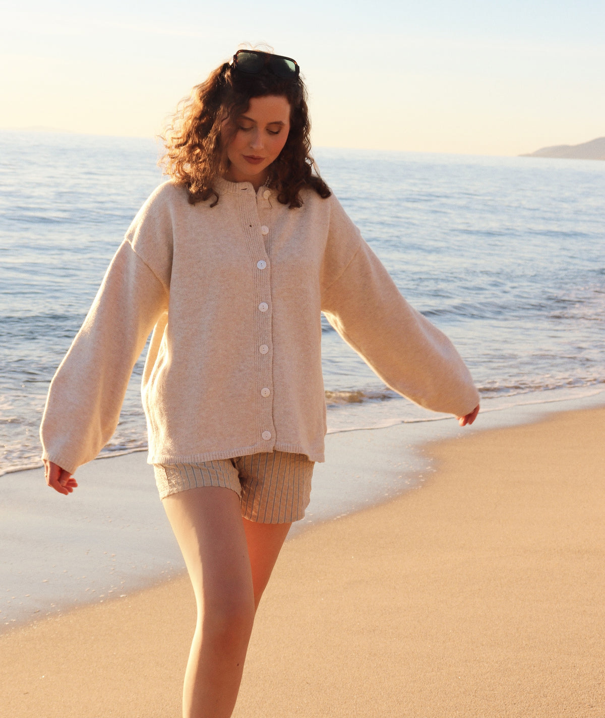 Woman in a beige cardigan and shorts walking on a sandy beach with ocean in the background