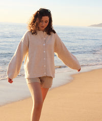 Woman in a beige cardigan and shorts walking on a sandy beach with ocean in the background