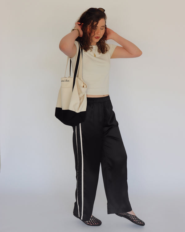 A woman stands against a plain light background wearing a cream crop top, black wide-leg pants with a white side stripe, black  flats, and carrying a black and cream tote bag.