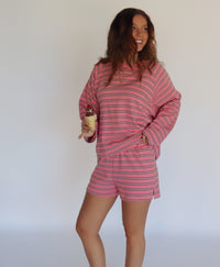 A woman modeling a pink and brown striped lounge set consisting of a long-sleeve top and matching shorts, posed against a plain white studio background.