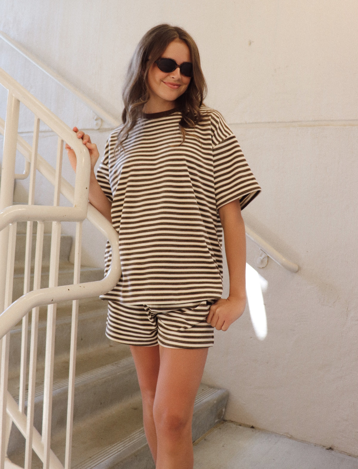 A woman wearing a matching brown and white striped lounge set consisting of a short-sleeve t-shirt and shorts, standing on a white staircase.