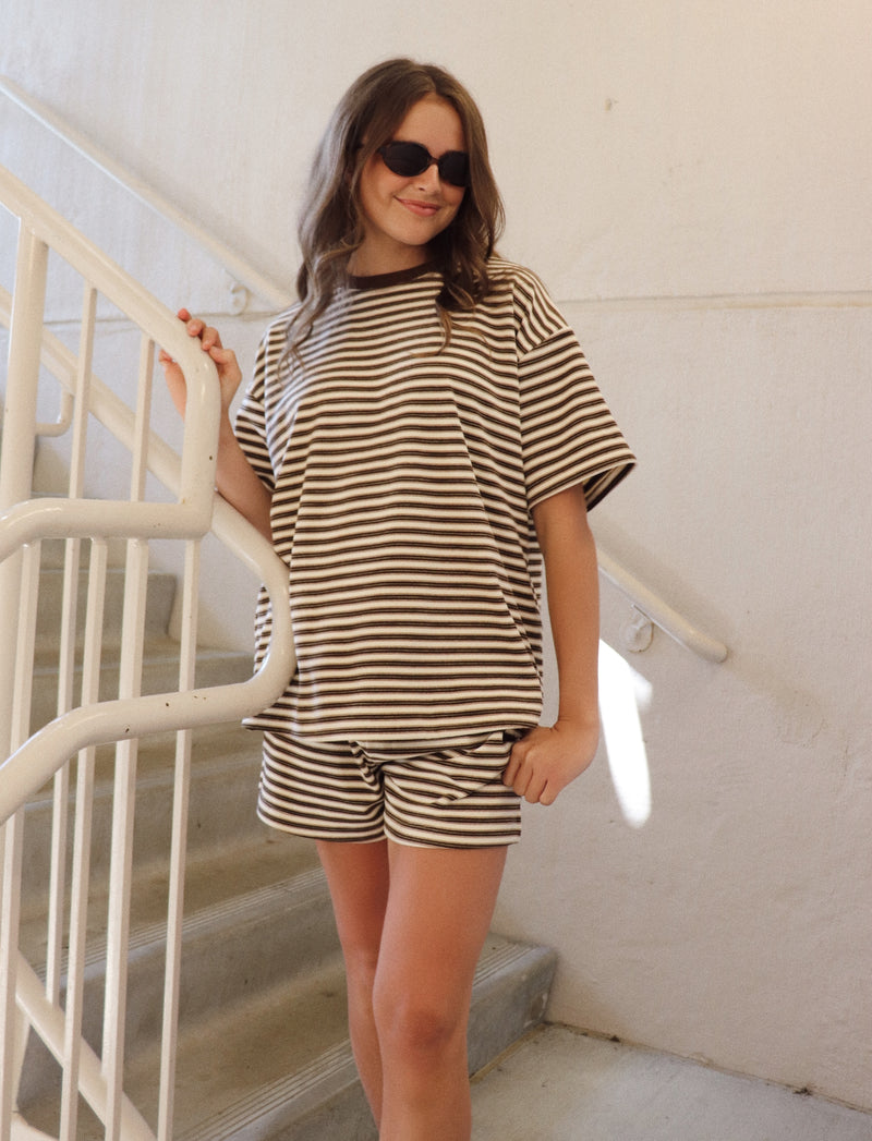 A woman wearing a matching brown and white striped lounge set consisting of a short-sleeve t-shirt and shorts, standing on a white staircase.