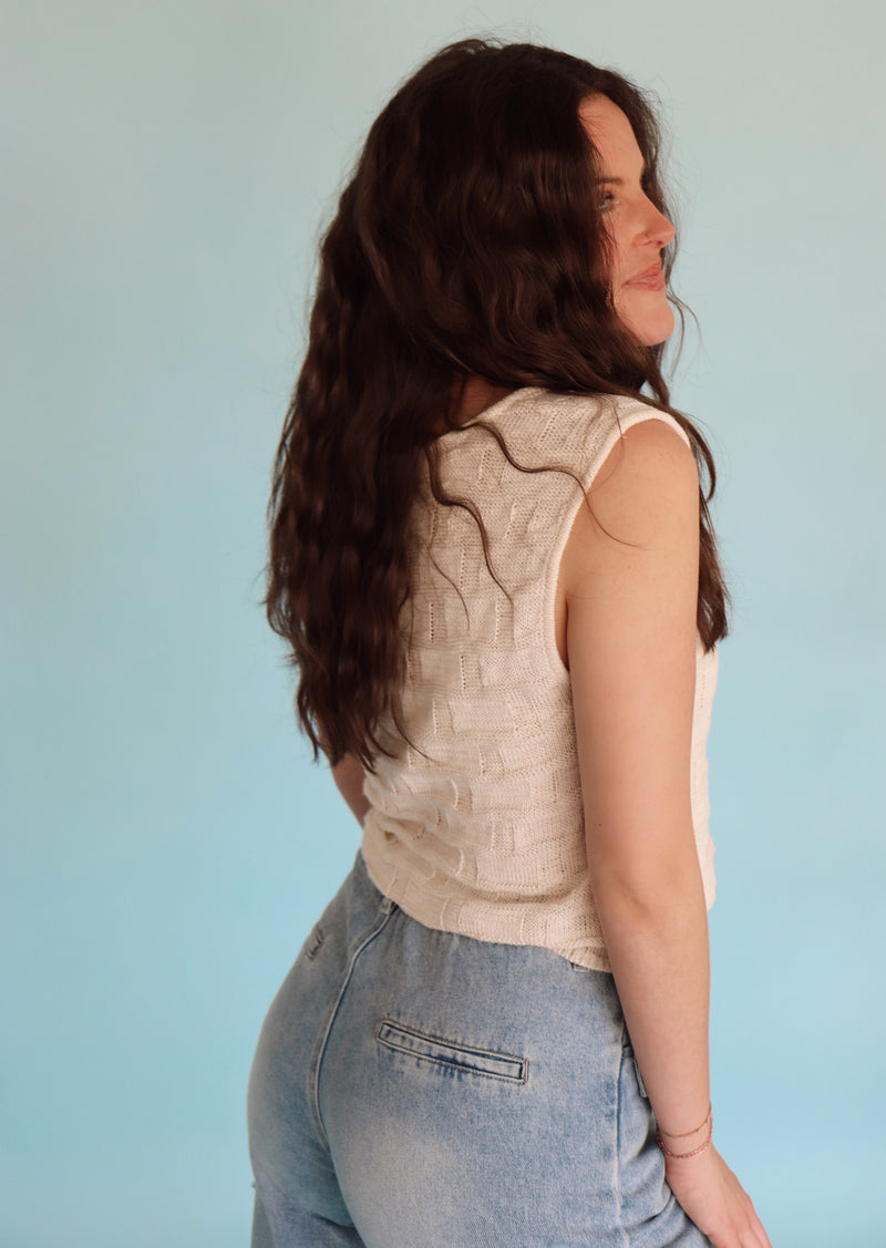 Woman wearing a cream vest and blue jeans against a light blue background