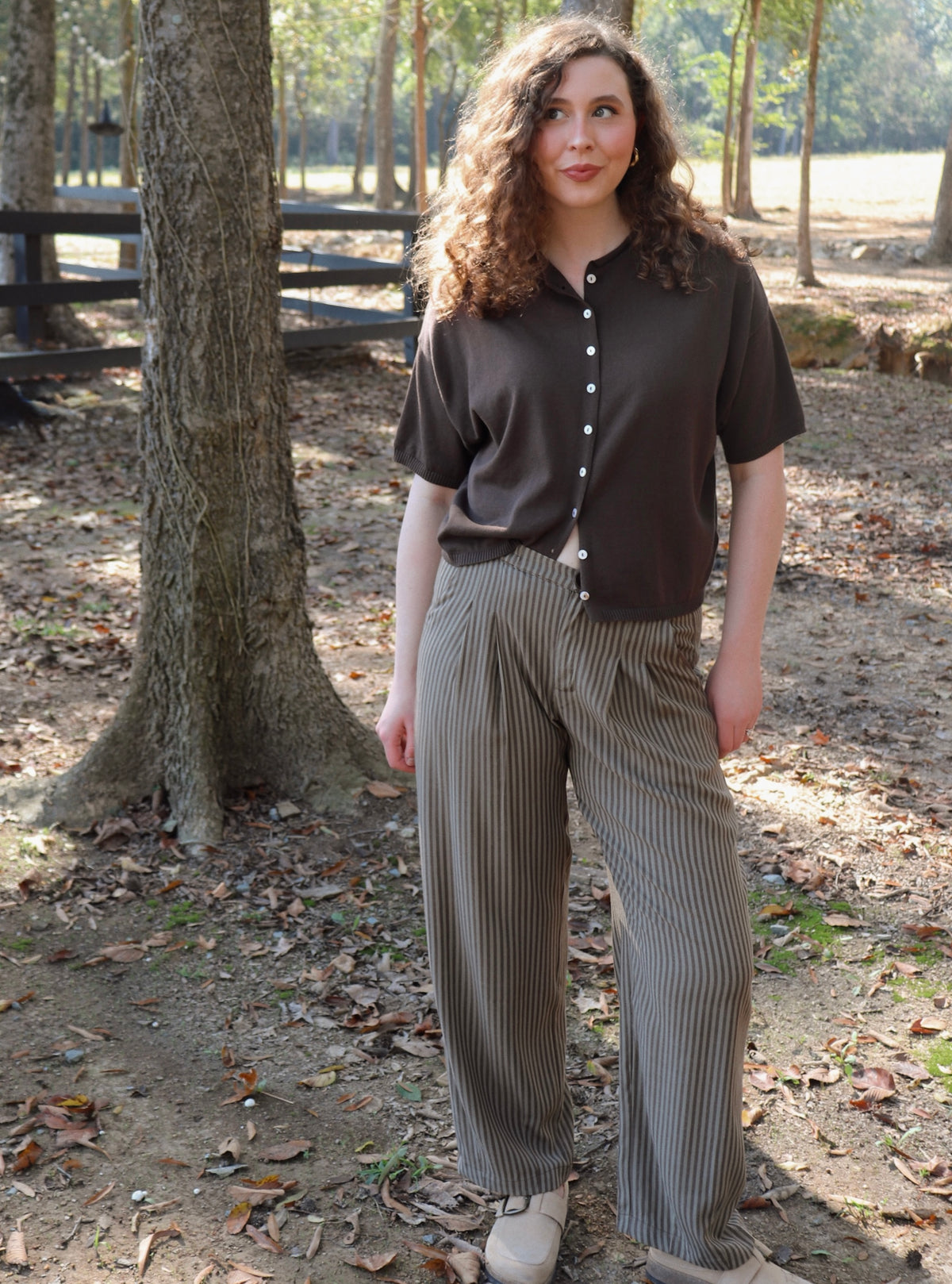 a woman wearing a chocolate brown short sleeve cardigan with brown striped pants stands in an outdoor setting