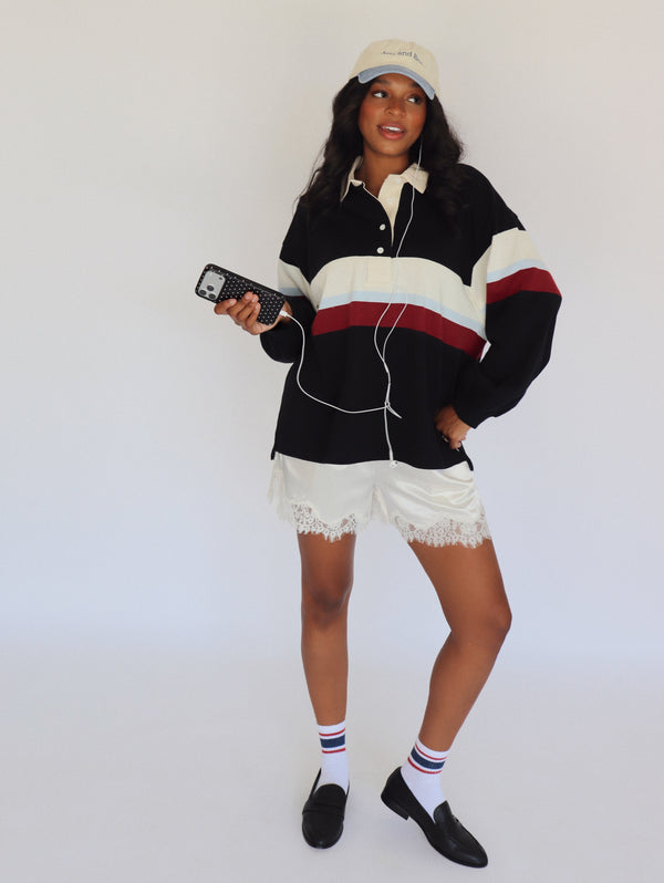 A woman modeling a black, white, and red striped rugby shirt with white shorts, white socks, black loafers, and a beige baseball cap, holding a phone against a plain white background.