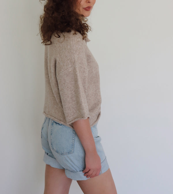A woman with curly brown hair wearing a light beige short-sleeve button-up knit top and light blue denim shorts, standing against a plain white wall.