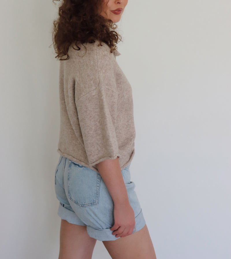 A woman with curly brown hair wearing a light beige short-sleeve button-up knit top and light blue denim shorts, standing against a plain white wall.