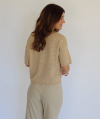 A woman modeling a beige short-sleeve button-front top with a subtle dotted texture, paired with beige and white striped pants, against a plain light background.