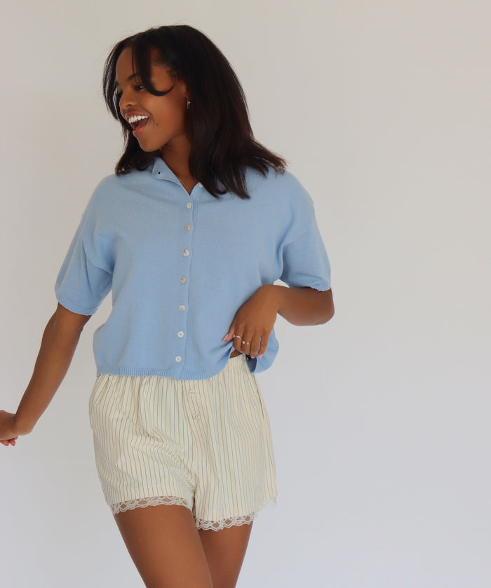 Woman wearing a blue short sleeve cardigan and cream shorts against a white background