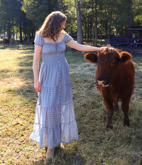 A woman wearing a light blue and white gingham tiered maxi dress stands in a grassy field next to a brown cow, touching its head. Trees and a fence are visible in the background.