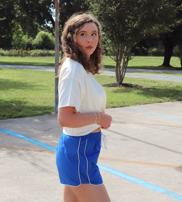 Woman in a white top and blue shorts standing on a concrete surface with grass and trees in the background