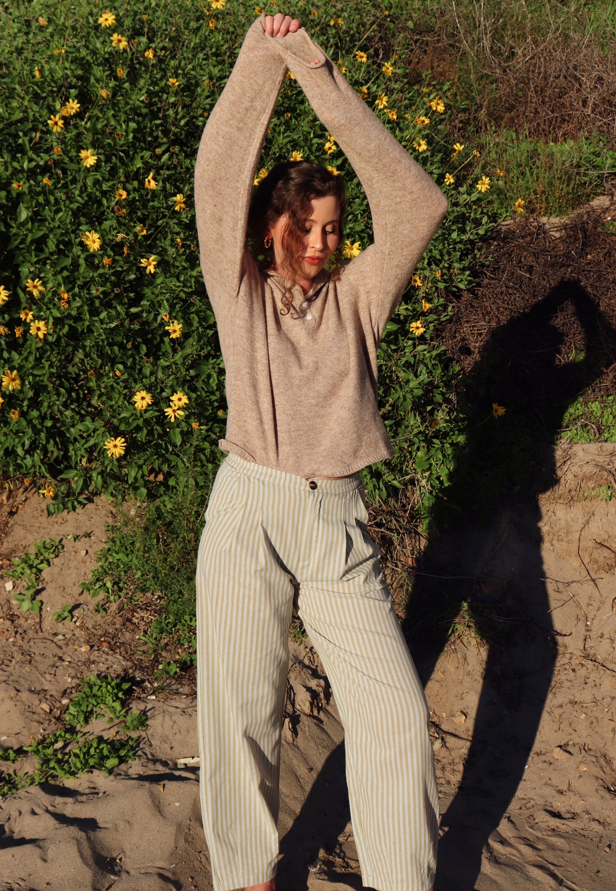 Person outdoors with greenery and flowers in the background wearing a brown top and striped pants.