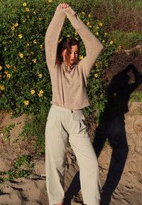 Person outdoors with greenery and flowers in the background wearing a brown top and striped pants.