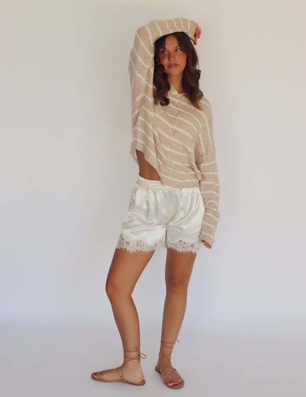 A woman modeling a beige and white striped knit long-sleeve top paired with white satin shorts featuring lace trim, standing against a plain white studio background.
