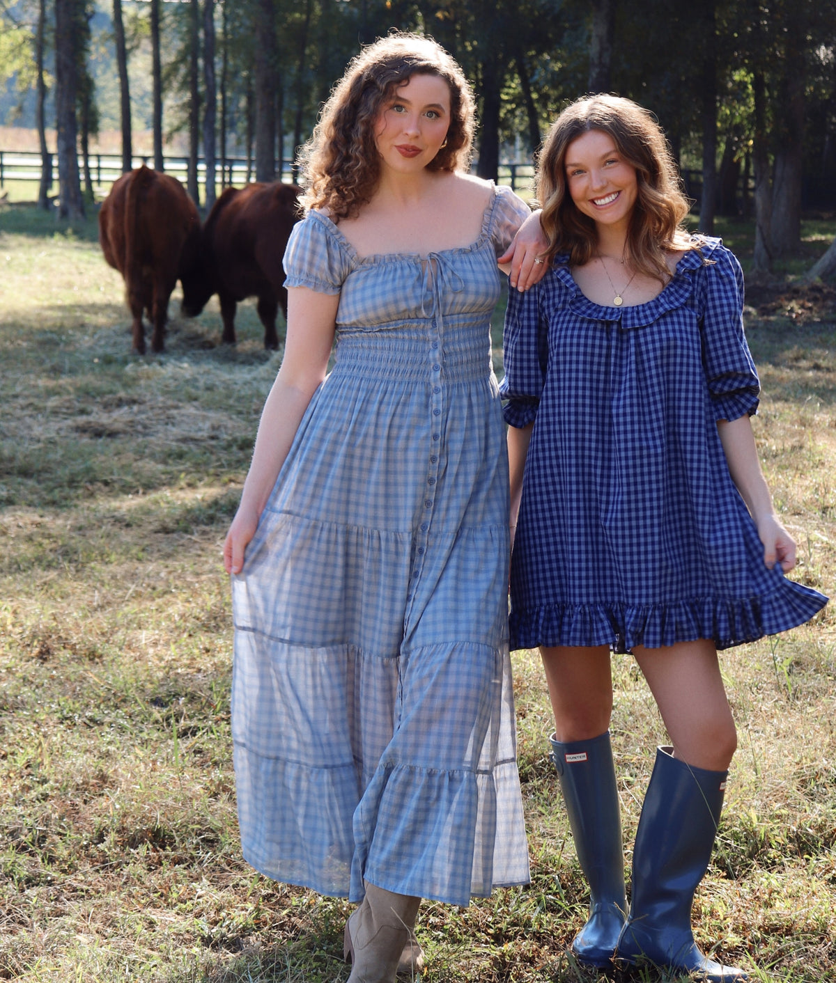 Two women standing outdoors in a grassy field with trees and cows in the background, wearing gingham dresses. The woman on the left wears a light blue and white checkered midi dress, and the woman on the right wears a dark blue and light blue checkered mini dress with blue rain boots.