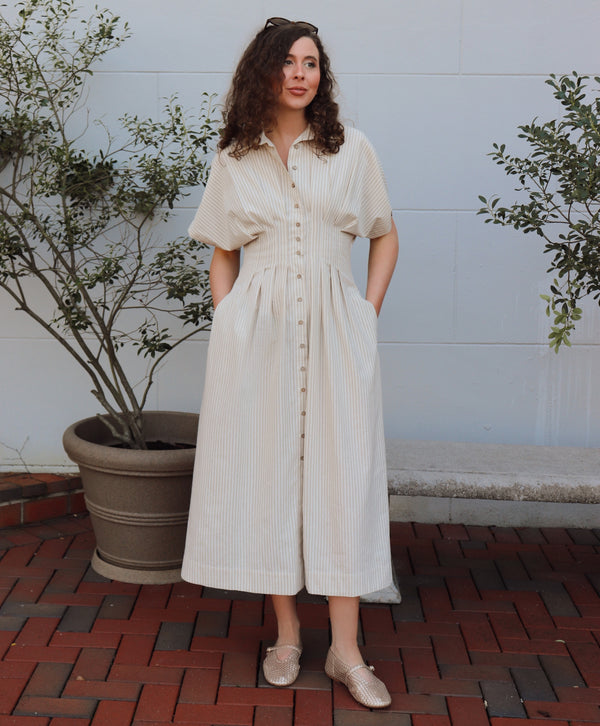 Woman in a taupe and cream striped  dress standing outdoors with plants and a bench in the background