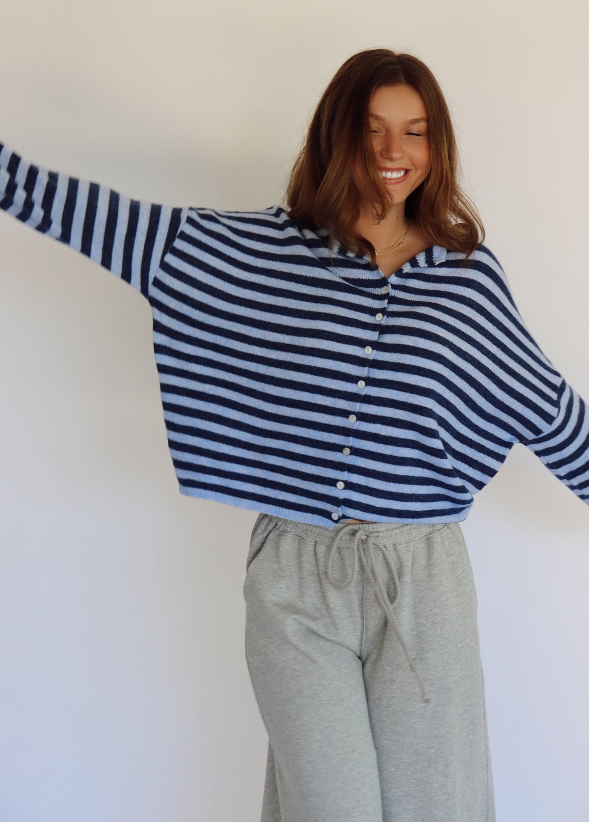 A woman wearing a navy and light blue horizontally striped long-sleeve button-up cardigan and light gray sweatpants, standing against a plain off-white wall.