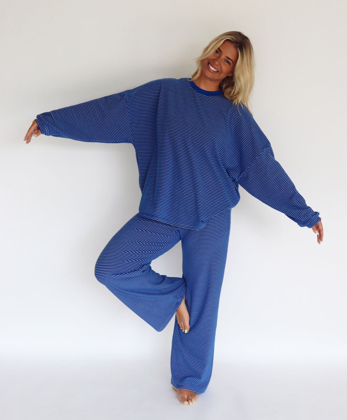 A woman modeling a matching blue striped lounge set consisting of a long-sleeve top and wide-leg pants, standing against a plain white studio background.