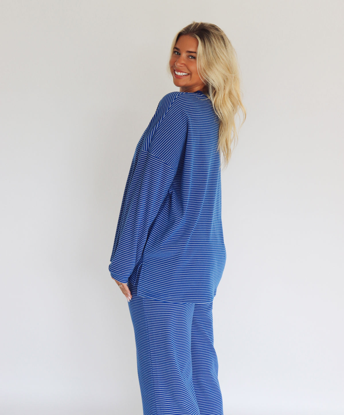 A woman modeling a matching blue striped lounge set consisting of a long-sleeve top and wide-leg pants, standing against a plain white studio background.