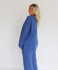 A woman modeling a matching blue striped lounge set consisting of a long-sleeve top and wide-leg pants, standing against a plain white studio background.