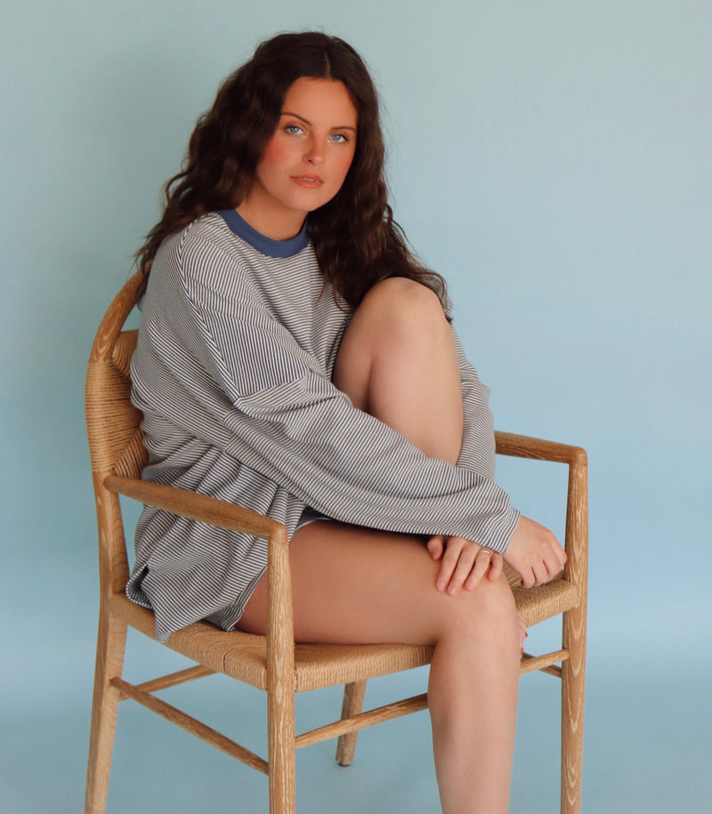 Woman sitting on a wooden chair wearing a blue striped top and shorts against a light blue background