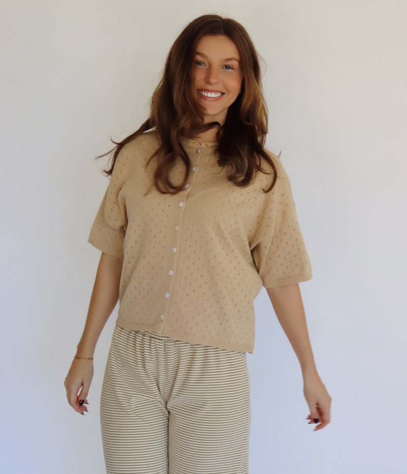 A woman modeling a beige short-sleeve button-front top with a subtle dotted texture, paired with beige and white striped pants, against a plain light background.