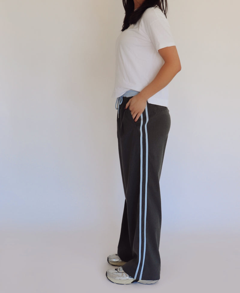 Person wearing dark gray pants with blue stripes and a white shirt against a plain background