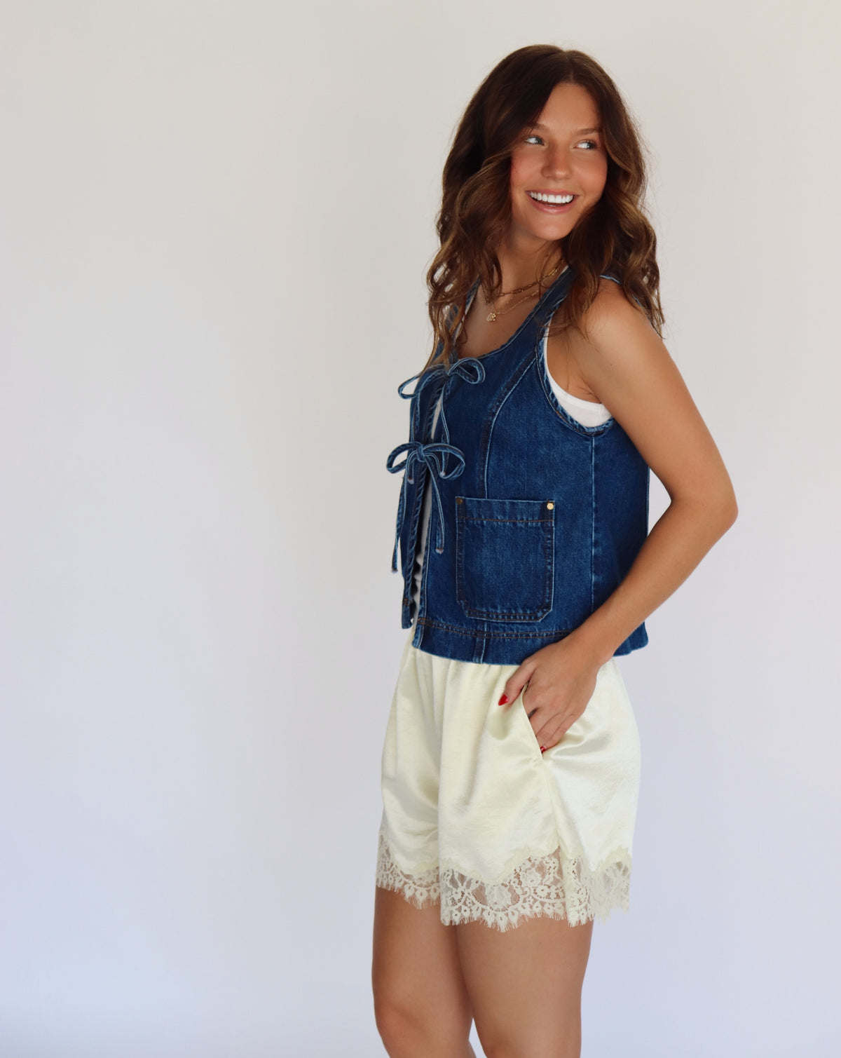 A woman modeling a sleeveless blue denim vest with front tie details over a white top and yellow satin shorts with lace trim, set against a plain background.