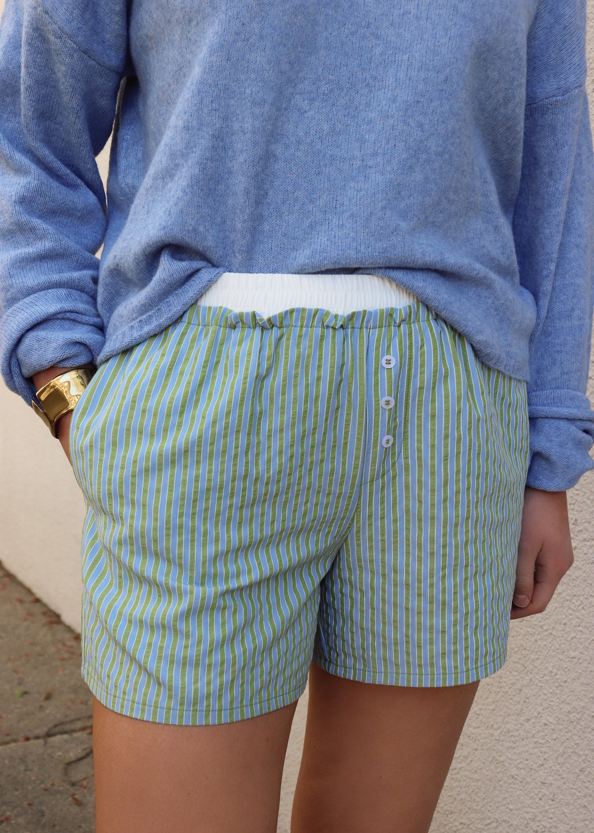 Woman wearing a blue sweater and green and blue striped shorts against a gray wall.