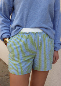 Woman wearing a blue sweater and green and blue striped shorts against a gray wall.