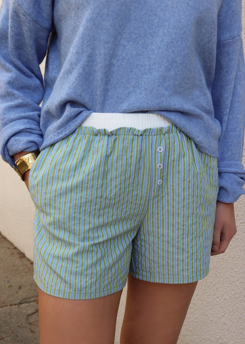 Woman wearing a blue sweater and green and blue striped shorts against a gray wall.