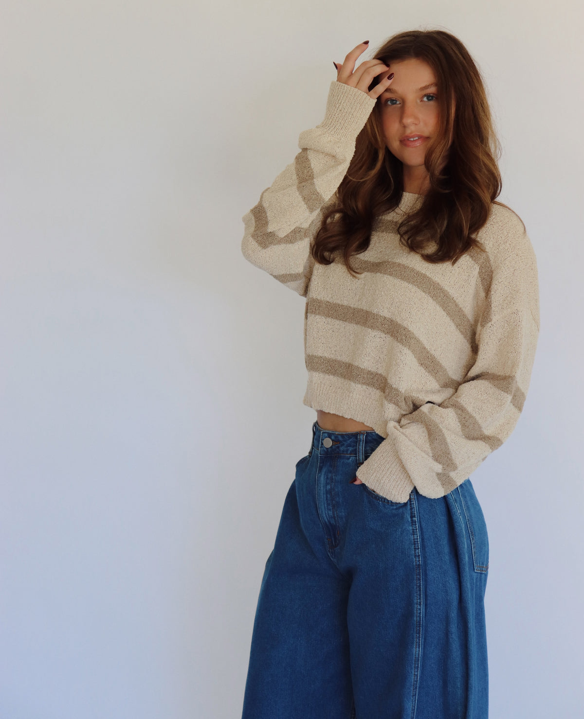 A woman wearing a taupe and tan striped knit sweater and high-waisted blue jeans, posing against a plain light gray wall.