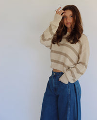 A woman wearing a taupe and tan striped knit sweater and high-waisted blue jeans, posing against a plain light gray wall.
