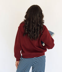 A woman viewed from behind wearing a burgundy crewneck sweatshirt and blue-and-navy striped pants, holding a book against a plain white background.