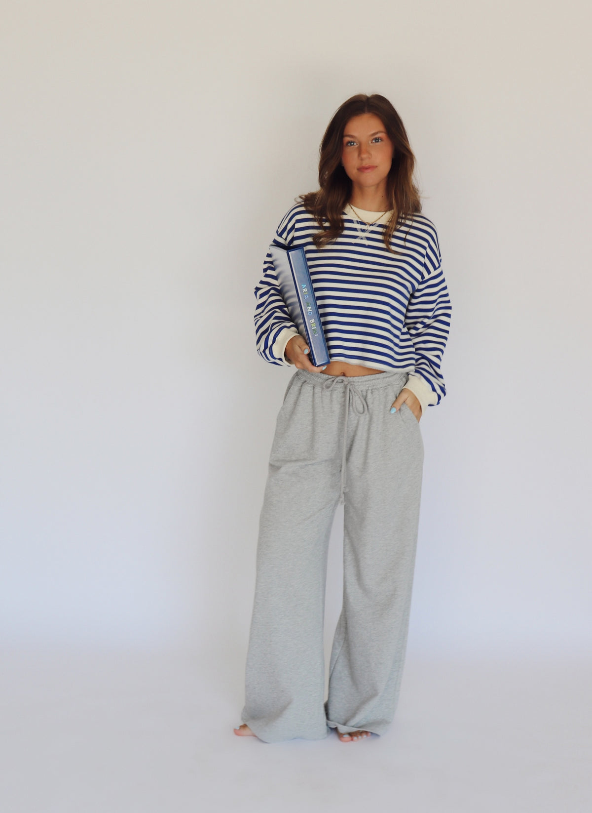 A woman stands against a plain white studio background wearing a blue and off white striped long-sleeve crop top, light gray wide-leg sweatpants, and holding a blue book.