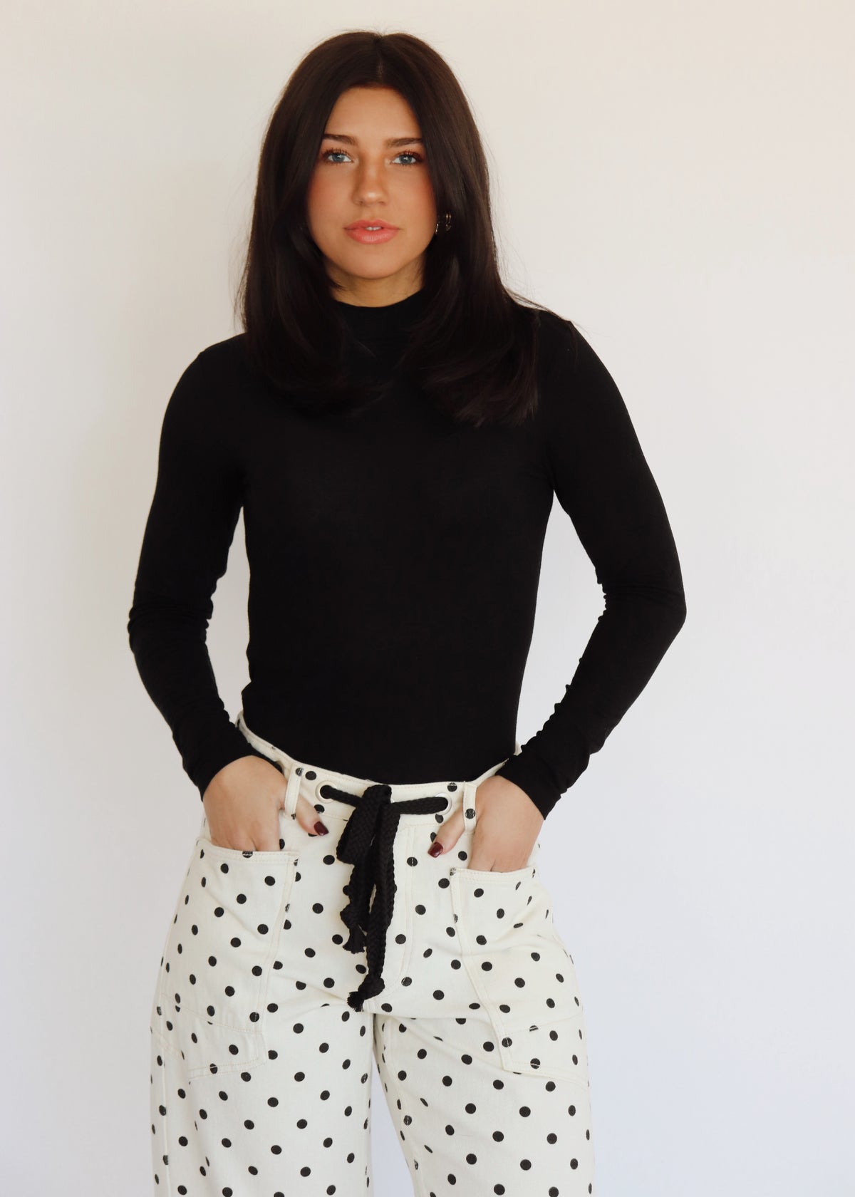 A woman in a black turtleneck and white barrel jeans with black polka dots on them, in front of a white background