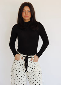 A woman in a black turtleneck and white barrel jeans with black polka dots on them, in front of a white background
