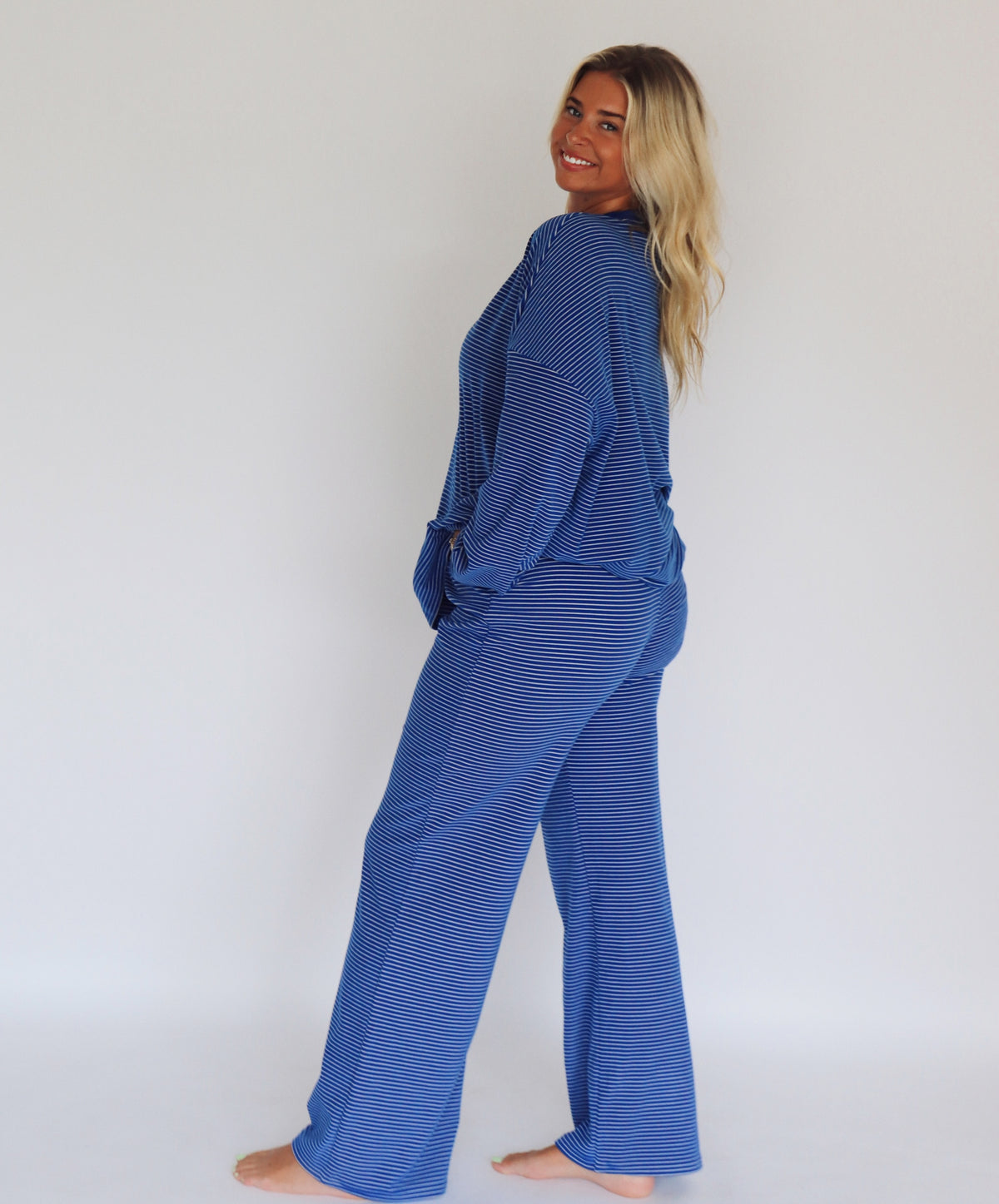 A woman modeling a matching blue striped lounge set consisting of a long-sleeve top and wide-leg pants, standing against a plain white studio background.