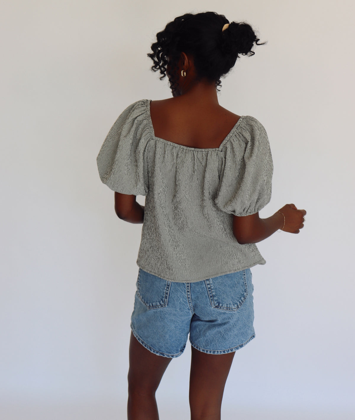 Woman wearing a black and white gingham blouse with puffed sleeves and denim shorts against a white background