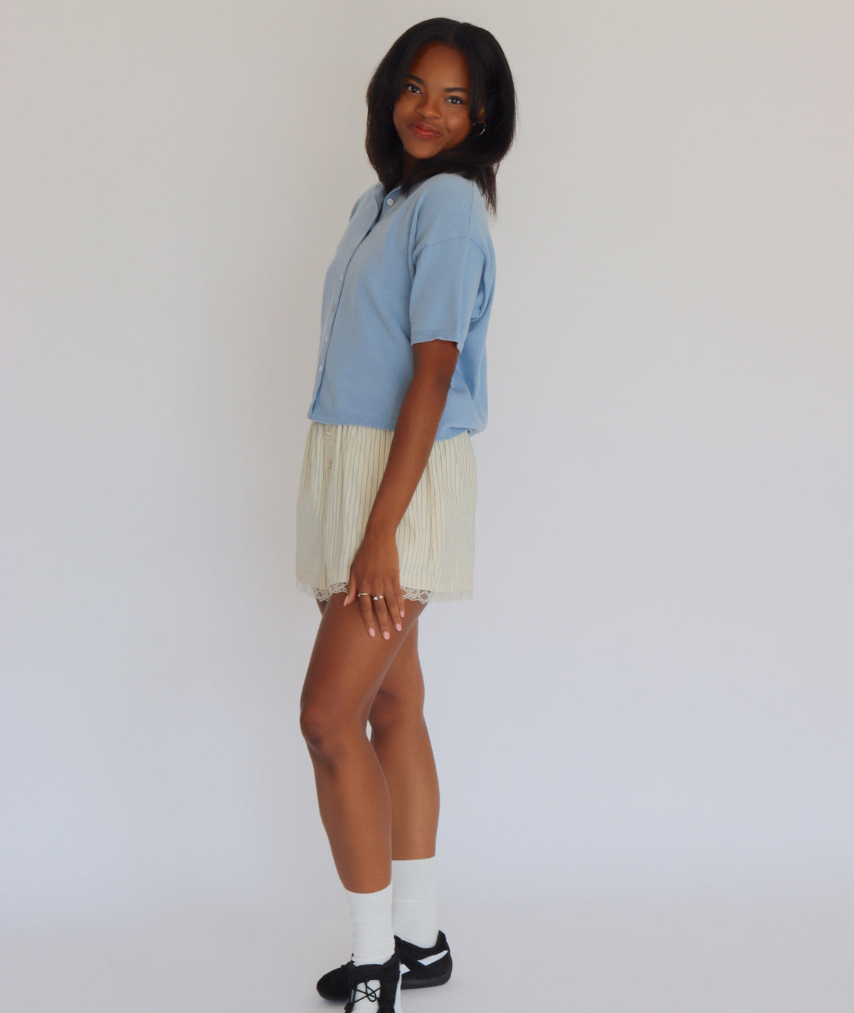 Woman wearing a blue short sleeve cardigan and cream shorts against a white background
