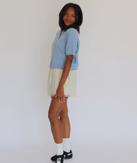 Woman wearing a blue short sleeve cardigan and cream shorts against a white background