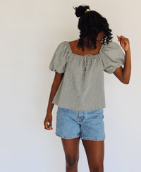 Woman wearing a black and white gingham blouse with puffed sleeves and denim shorts against a white background