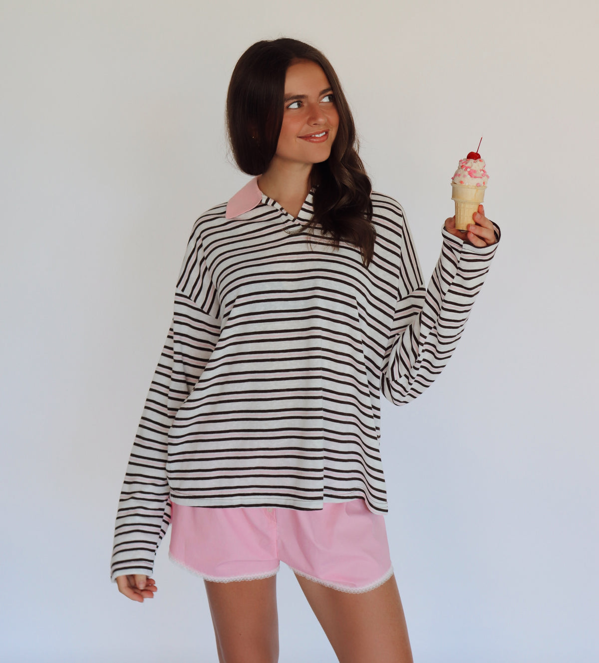 A woman modeling a brown, pink and white horizontally striped long-sleeve polo shirt with a pink collar, paired with light pink shorts, while holding a pink-frosted ice cream cone against a plain white background.