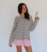 A woman modeling a brown, pink and white horizontally striped long-sleeve polo shirt with a pink collar, paired with light pink shorts, while holding a pink-frosted ice cream cone against a plain white background.