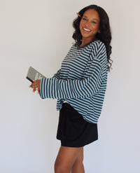 A woman modeling a long-sleeve blue striped top with shorts, holding a black and white notebook bag against a plain white background.