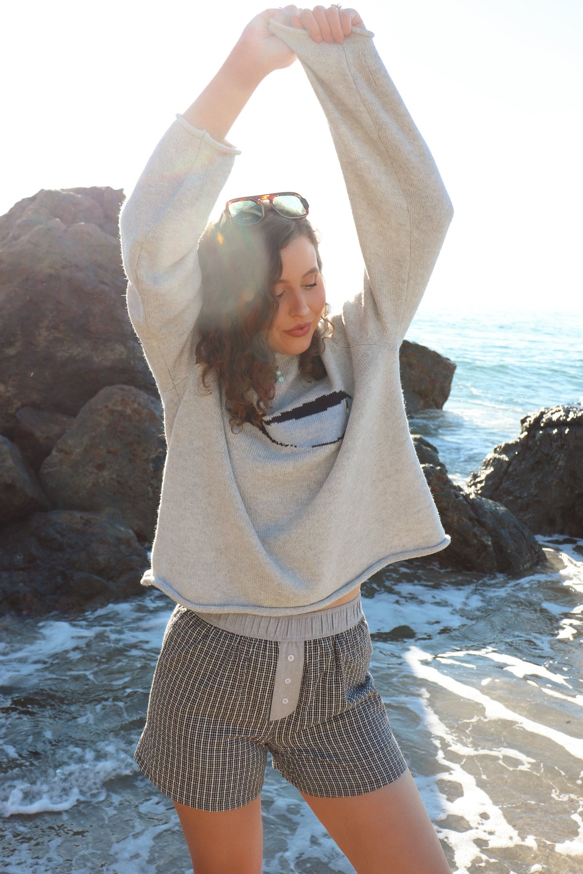 Woman outdoors by a rocky coastline, wearing a gray fish sweater and black shorts. 