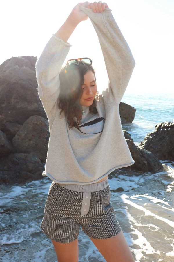 Woman outdoors by a rocky coastline, wearing a gray fish sweater and black shorts. 
