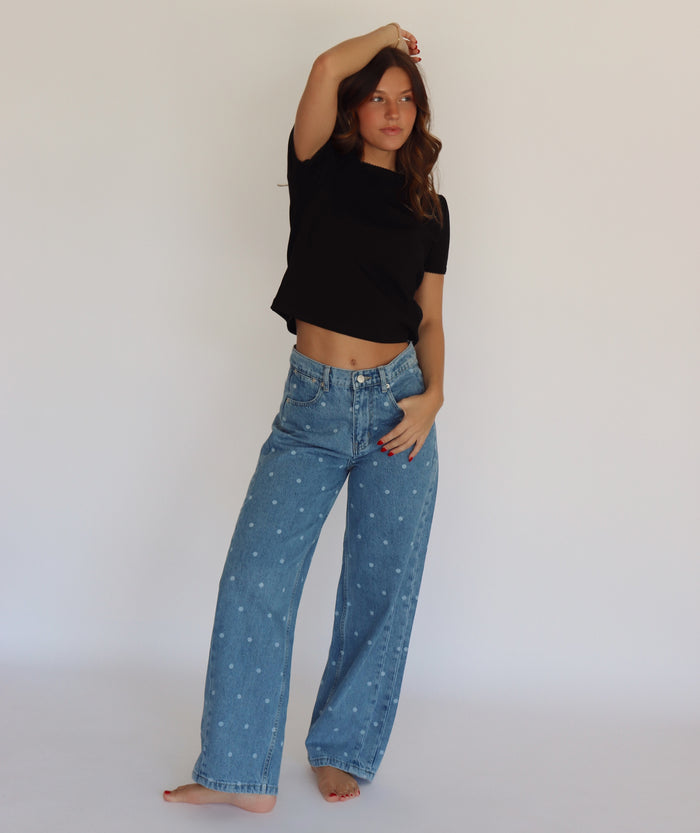 A woman modeling a black short-sleeve crop top and light blue wide-leg denim jeans with small white polka dots, standing against a plain white studio background.